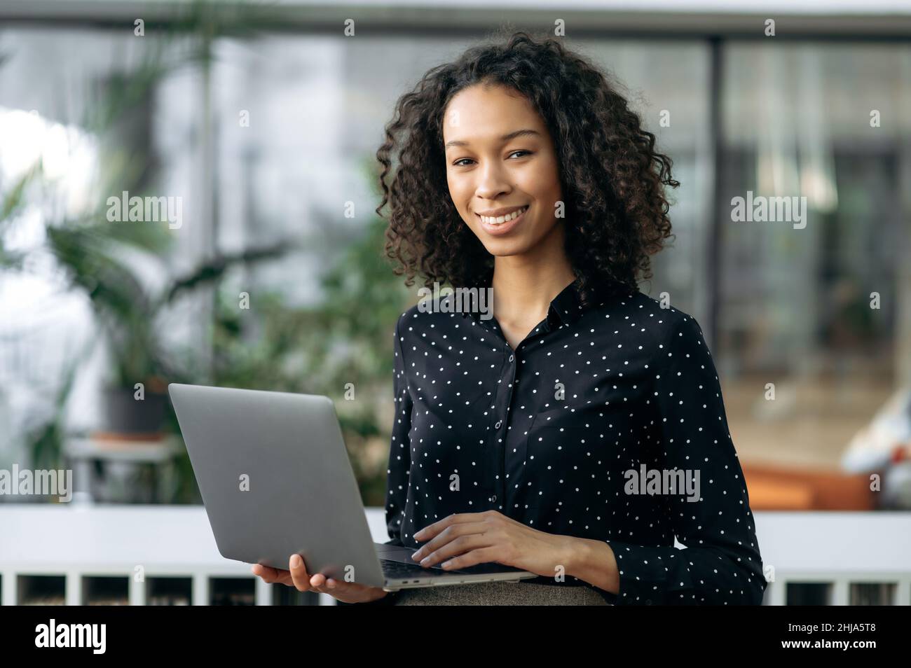Porträt von lockig attraktiven positiven afroamerikanischen angenehme junge Geschäftsfrau, Freiberufler, Immobilienmakler oder Manager, mit offenem Laptop im Büro, Blick auf die Kamera, lächelnd freundlich Stockfoto