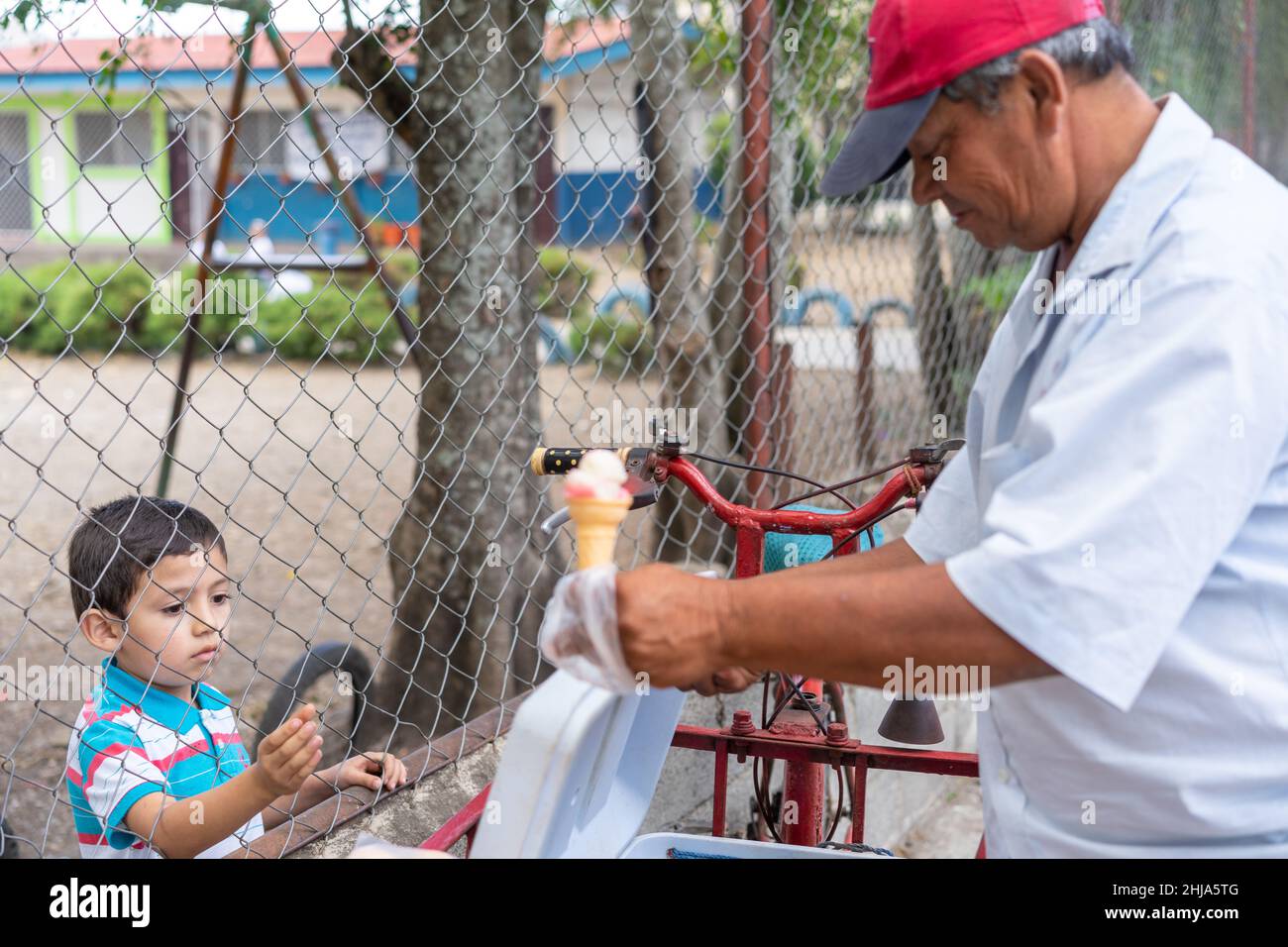 Ein Straßenhändler in der Nähe einer Grundschule übergibt einem Jungen in Jinotega, Nicaragua, einen Eiskegel durch einen Kettenzaun. Stockfoto