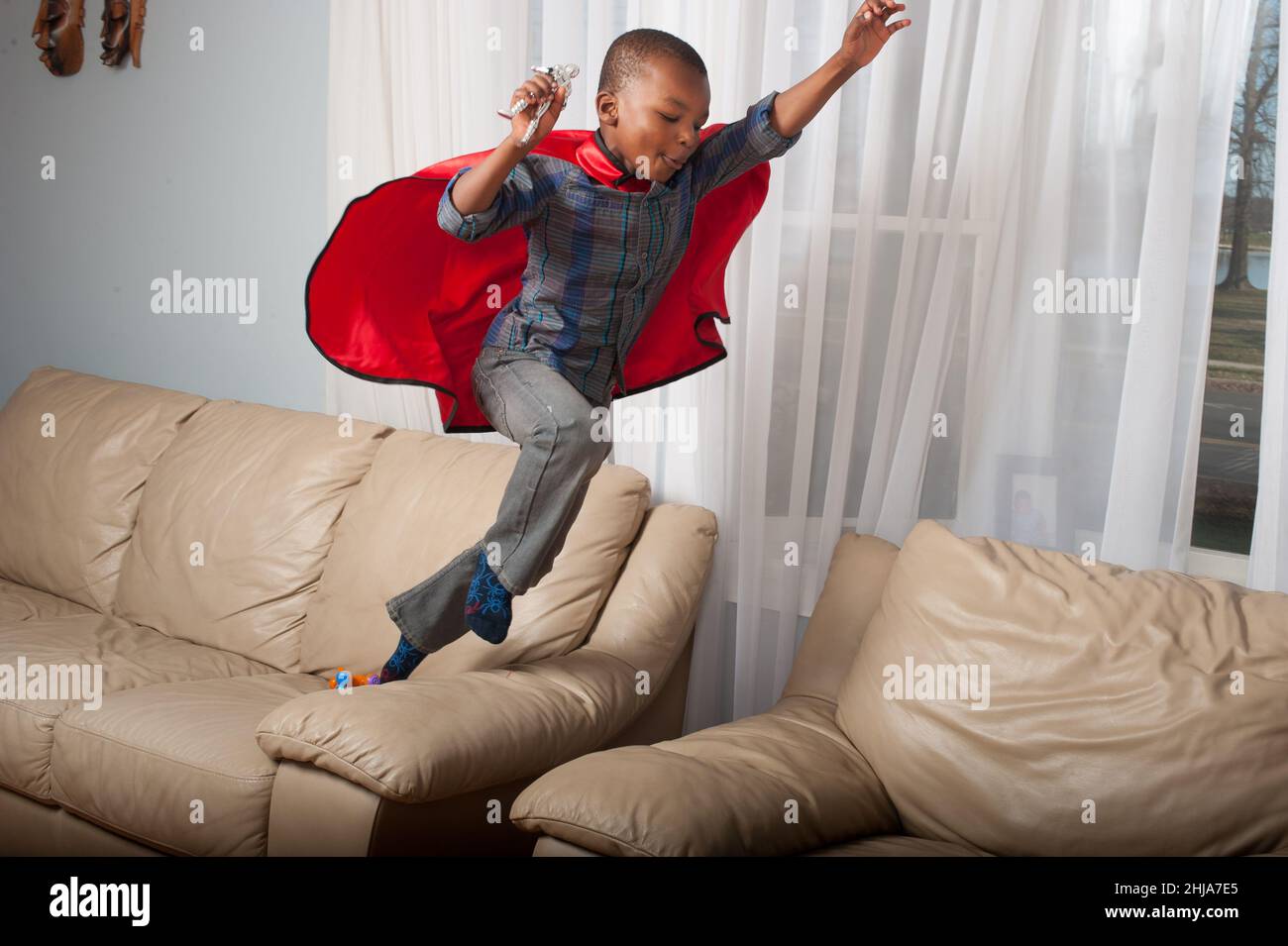 4-jähriger Junge mit Superhelden-Umhang, der von der Couch auf den Stuhl springt und Action-Figur hält Stockfoto