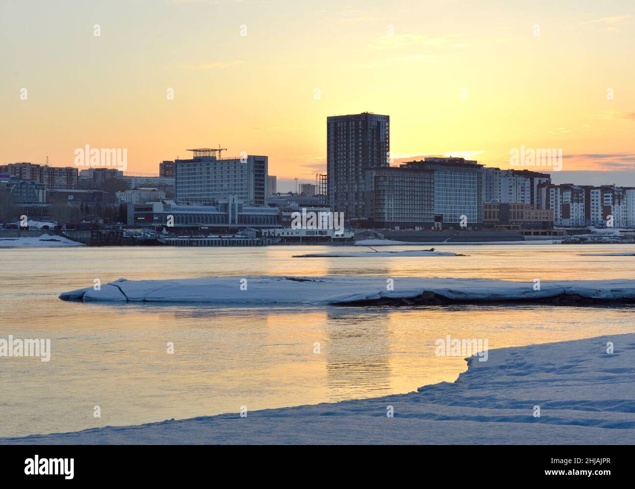 Die Obi-Küste im Morgengrauen, Schnee und Eis am Ufer im Frühling, hohe moderne Häuser am Horizont, Spiegelungen im Wasser, goldener Himmel. Russland, Sibirien, Stockfoto