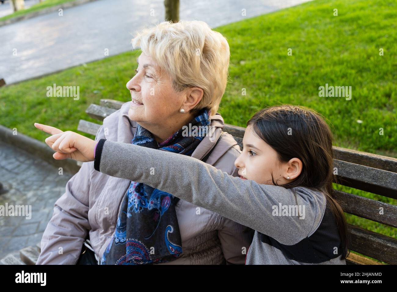 Kleines Mädchen zeigt mit Finger und Großmutter, die in diese Richtung schaut. Ältere Frau, die mit der Enkelin auf der Parkbank sitzt. Neugierde, Entdeckung Stockfoto