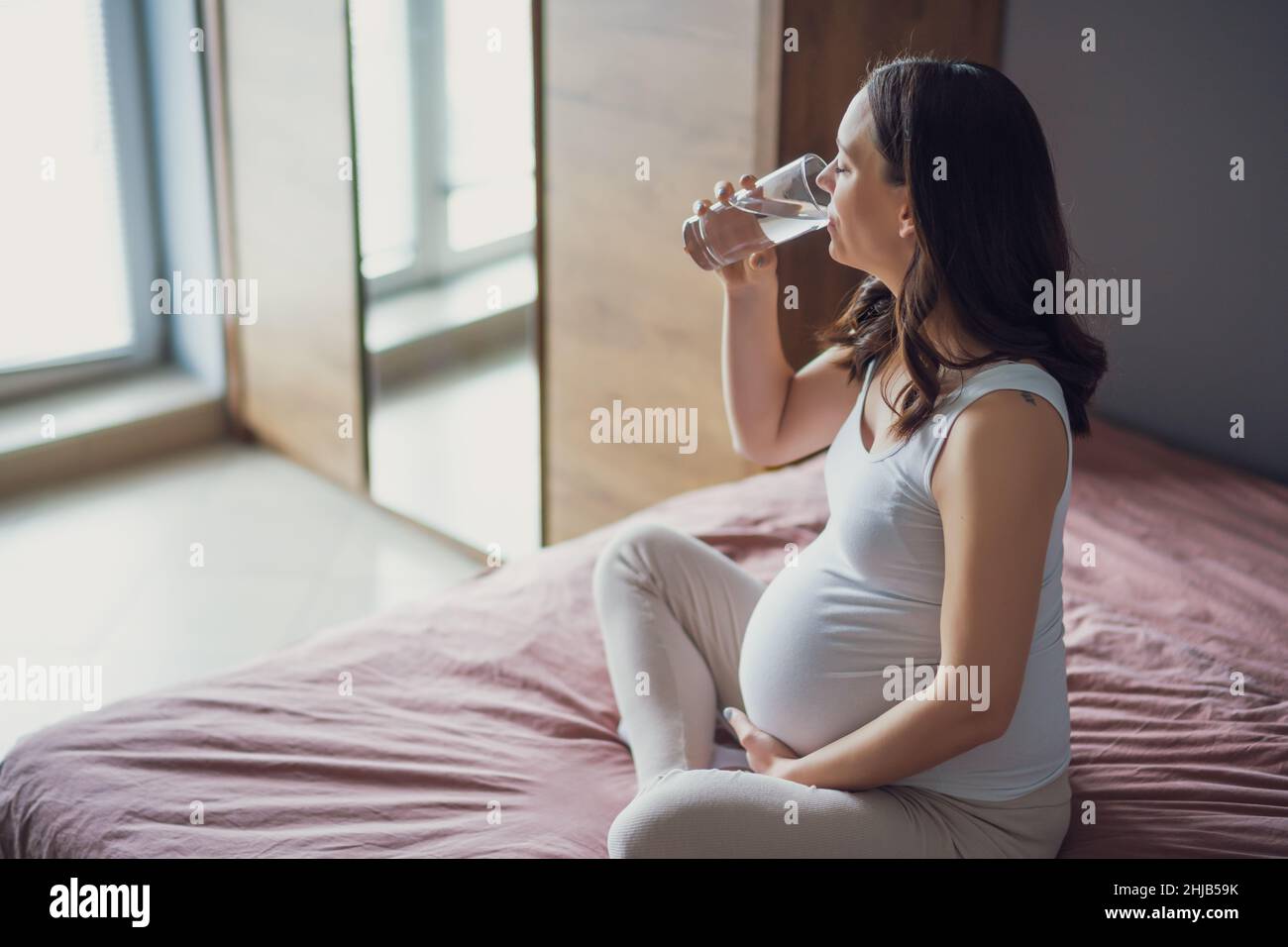 Schwangerin entspannt sich zu Hause. Sie sitzt auf dem Bett und trinkt Wasser. Stockfoto
