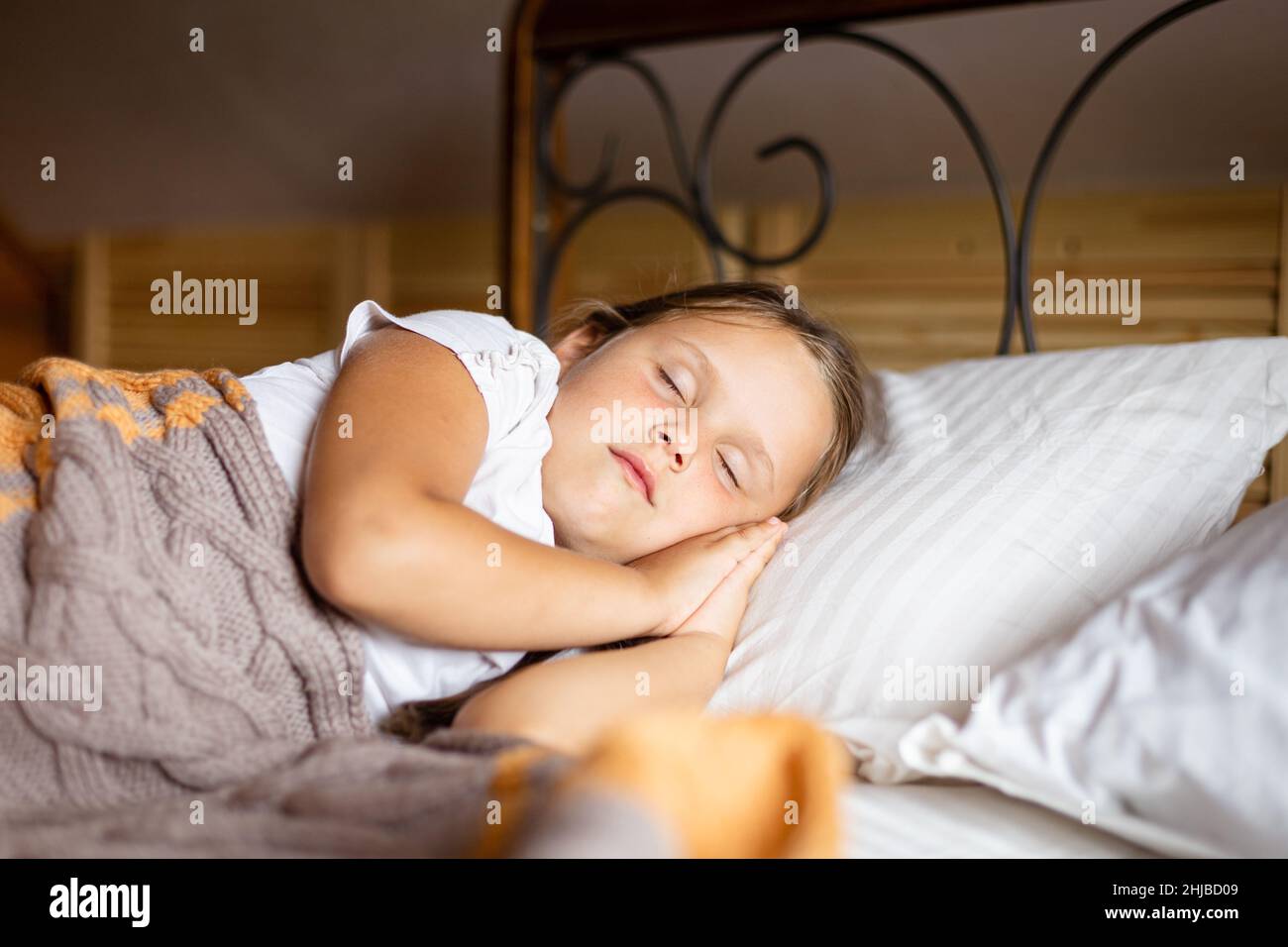 Nahaufnahme eines kleinen Mädchens, das auf einem großen Bett schläft, wobei beide Hände unter dem Kopf mit einer Decke bedeckt sind, die in einem Holzhaus Hauskleidung trägt. Schlafen Sie darin Stockfoto