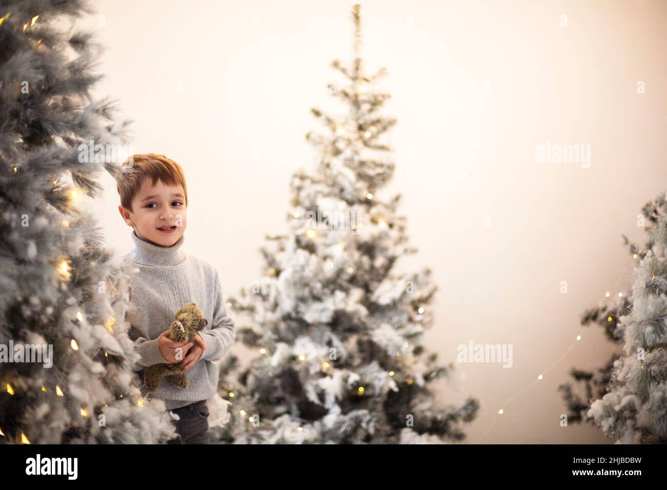 Weihnachtswunder. Niedlichen glücklichen kleinen Jungen mit Teddybär umgeben von schneebedeckten künstlichen Weihnachtsbäumen mit gelben funkelnden Lichtern geschmückt, sele Stockfoto