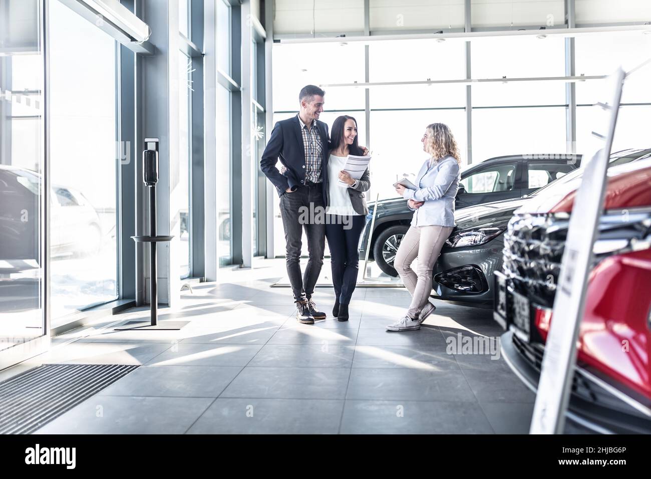Das junge Paar steht im Autohaus und diskutiert Optionen mit einer Verkäuferin, die sich gegen eines der Autos lehnt. Stockfoto