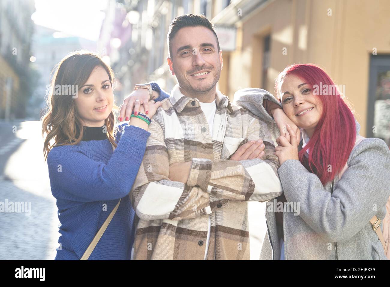 Positive Frauen lehnen sich mit gekreuzten Armen auf die Schultern eines Mannes und blicken lächelnd auf die Kamera, während sie an sonnigen Tagen auf der Straße der Stadt stehen Stockfoto