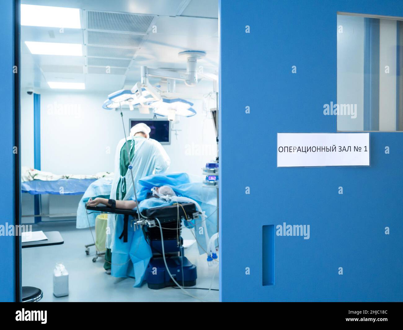 Selektiver Fokus auf die blaue Tür mit einem Schild in russischer Sprache: Operationssaal Nr. 1. Vor einem verschwommenen Hintergrund operieren Chirurgen auf dem Operationstisch. Stockfoto