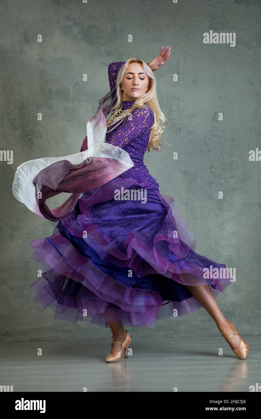 Blonde weibliche Ballroom-Tänzerin in fließender Tanzbewegung im lila Ballsaal vor grauem Hintergrund im Fotostudio Stockfoto