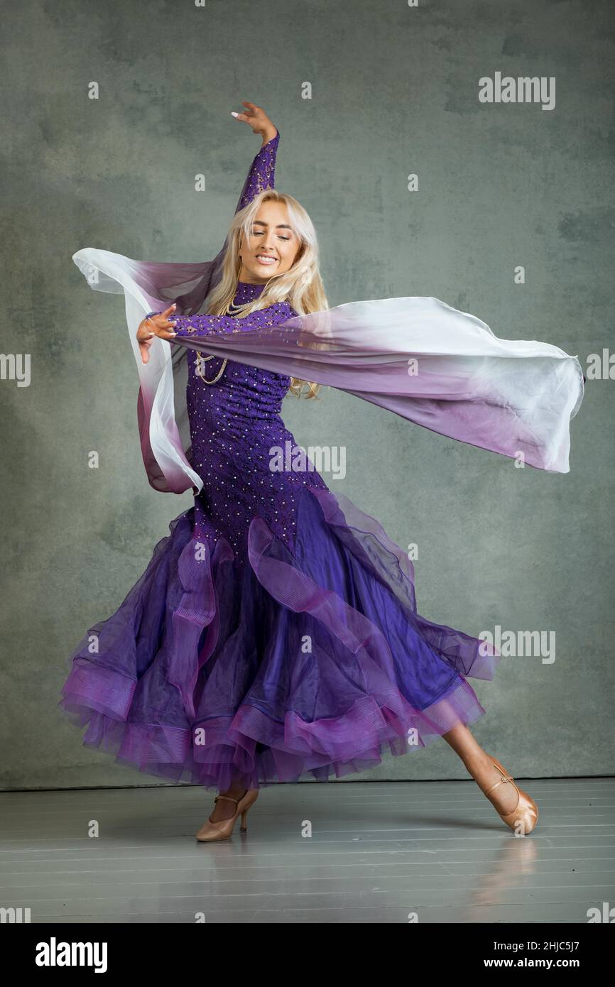 Blonde weibliche Ballroom-Tänzerin in fließender Tanzbewegung im lila Ballsaal vor grauem Hintergrund im Fotostudio Stockfoto