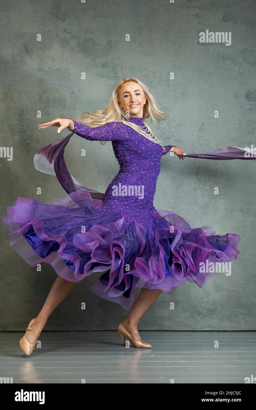 Blonde weibliche Ballroom-Tänzerin in fließender Tanzbewegung im lila Ballsaal vor grauem Hintergrund im Fotostudio Stockfoto