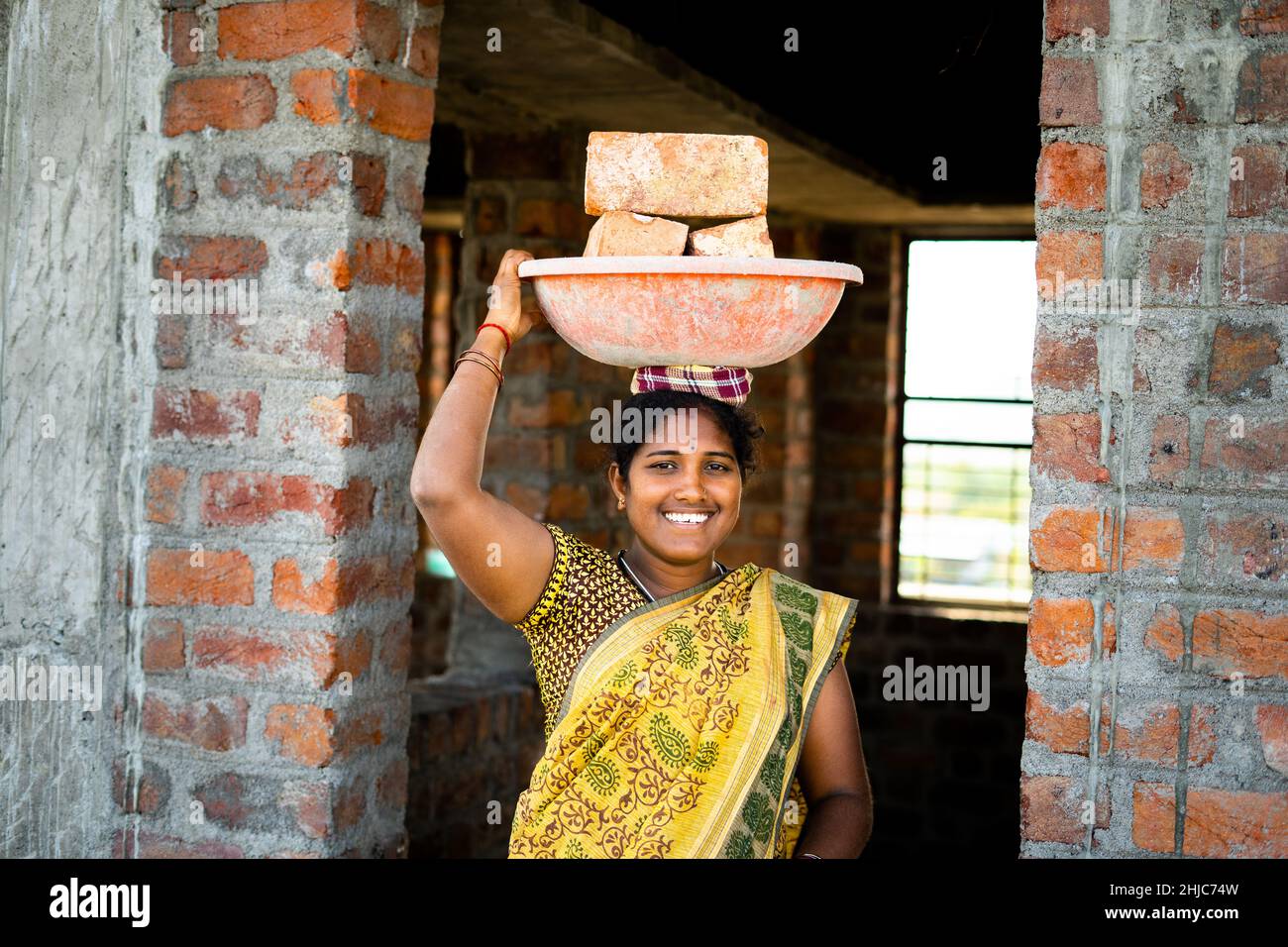 Fleißige, indisch lächelnde Frauen mit Ziegelsteinen auf dem Kopf, die die Kamera betrachten – Konzept des täglichen Wetteinsätzen, positiver emotionaler und weiblicher Ermächtigung. Stockfoto