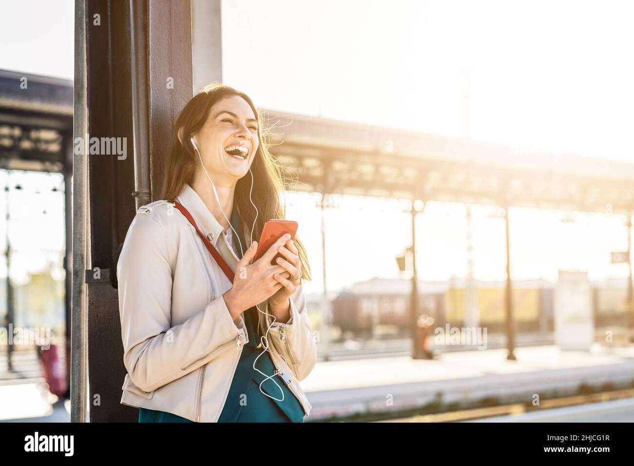 Frau in eleganten Kleidern wartet auf dem Bahnhofssteig mit Smartphone, um die Zeit zu verstreichen. Reise- und Geschäftsfrau-Konzept Stockfoto
