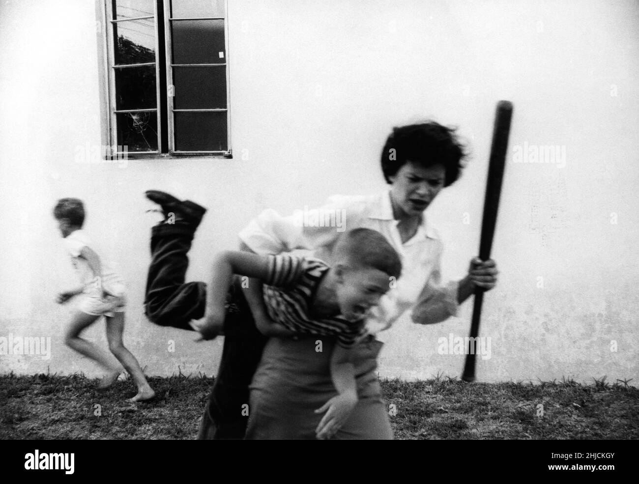 Wütend Mutter diszipliniert Sohn, nachdem er das Nachbarfenster mit einem Baseballspiel brach, während ein anderes Kind entkommt. Stockfoto