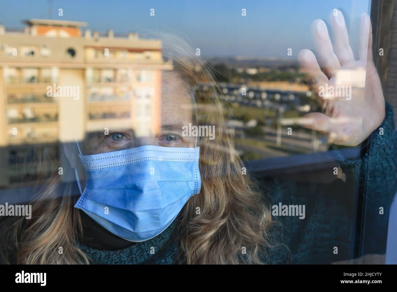 Durch das Coronavirus Covid-19 isolierte Frau schaut mit einer schützenden Gesichtsmaske durch das Glas ihres Hauses Stockfoto