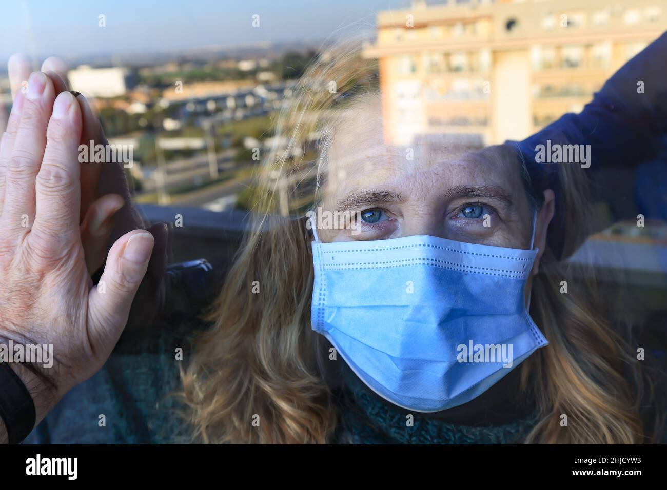 Durch das Coronavirus Covid-19 isolierte Frau schaut durch das Glas ihres Hauses zu ihrem Besucher und trägt eine schützende Gesichtsmaske Stockfoto