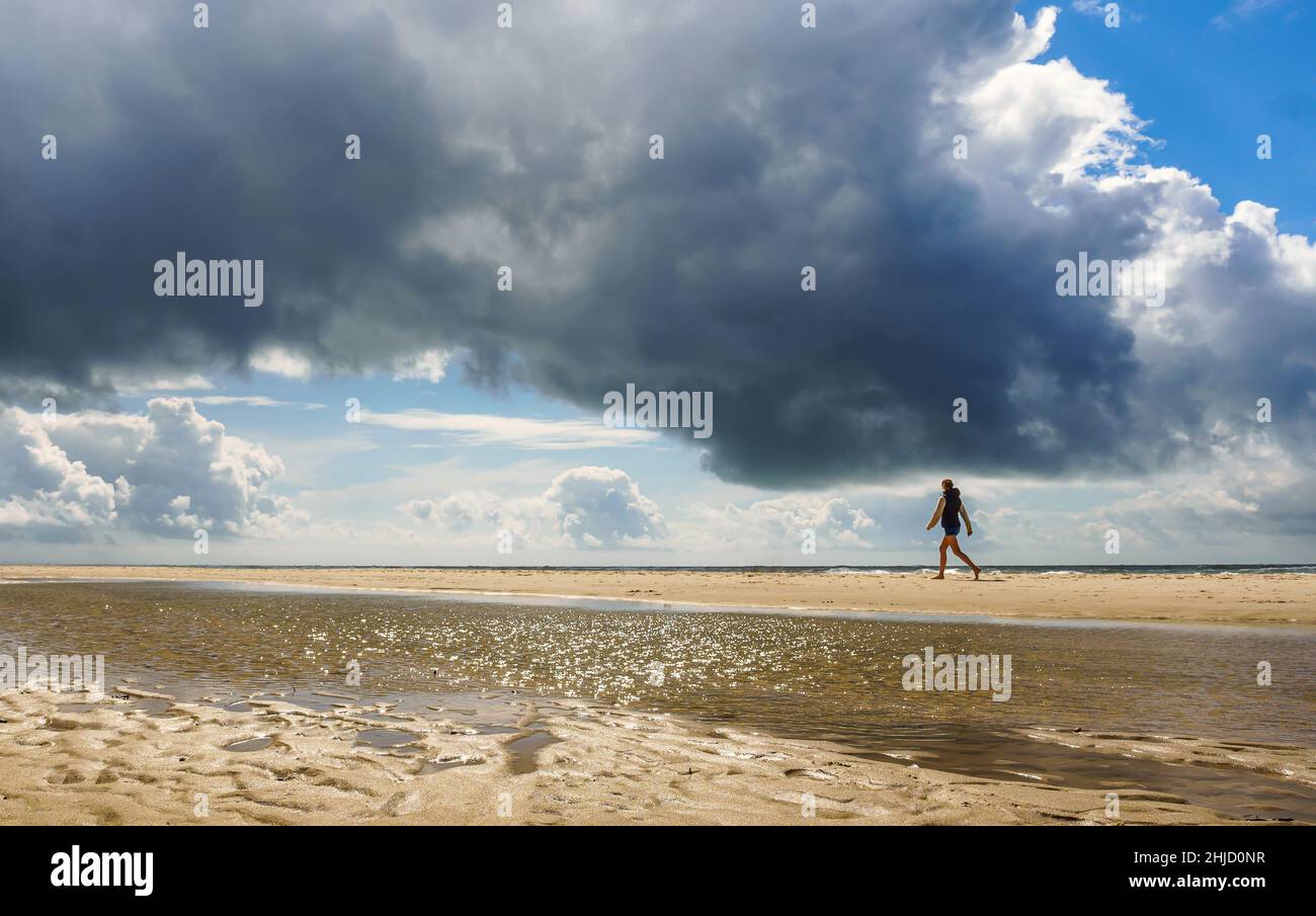 Eine junge Frau geht vor druckvoller Wolkenformation an einem menschenleeren Strand an dänischer Nordseeküste spazieren Stockfoto