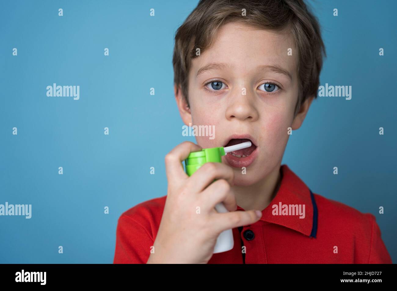 Kaukasischer Junge sprüht ein Aerosol in seinen Mund, um Halsschmerzen zu behandeln, Spray für die topische Anwendung. Aerosol Inhalator. Krankes Kind. Blauer Hintergrund. Kopierbereich Stockfoto