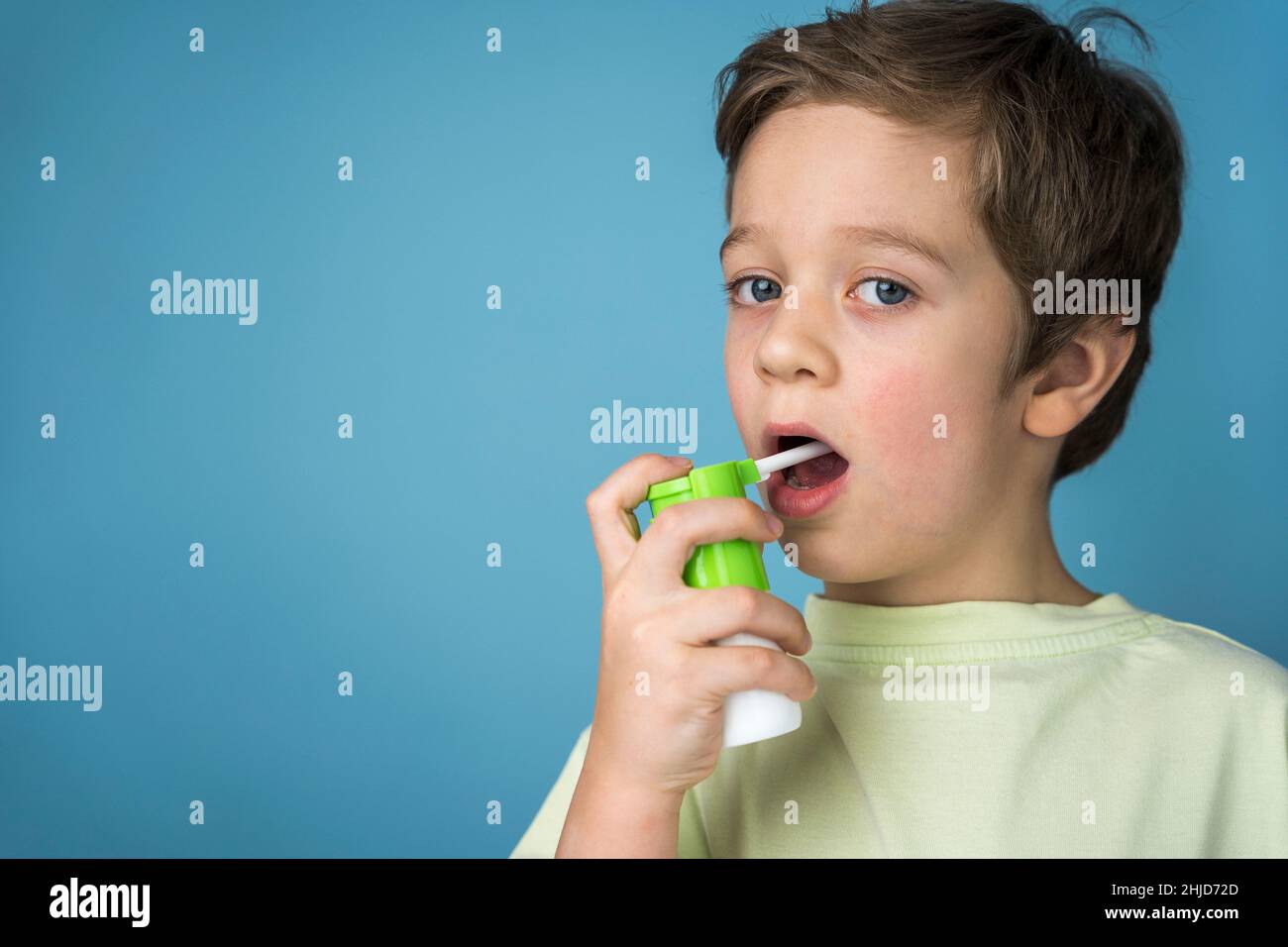 Ein süßer kaukasischer Junge in einem gelben T-Shirt sprüht ein Aerosol in seinen Mund, um Halsschmerzen zu behandeln, ein Spray für die topische Anwendung. Aerosol-Inhalator. Blaue Rückseite Stockfoto