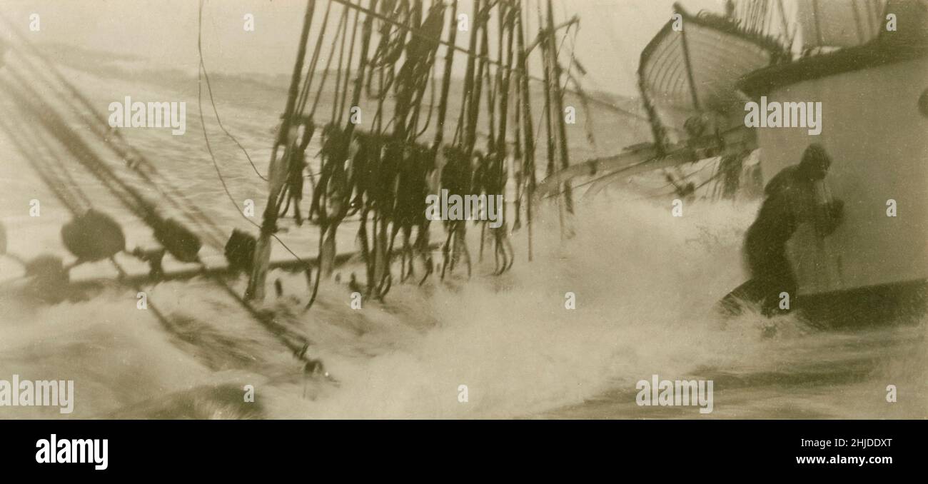 Raues Wetter auf See. Ein Seemann kämpft während eines Sturms auf See darum, sich aus dem Wasser zu halten. Das Segelschiff liegt auf einer Seite tief im Wasser und die Wellen überschwemmen das Deck. 1920s Stockfoto