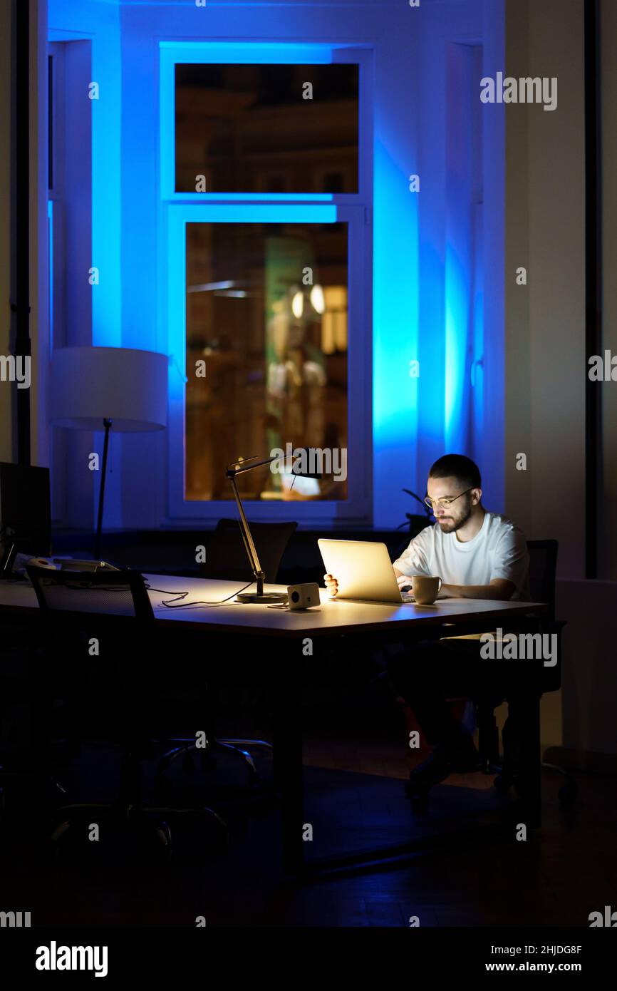 Nachdenklicher junger Geschäftsmann mit Brille arbeitet nachts auf dem Laptop und bereitet sich auf die Startup-Präsentation vor Stockfoto