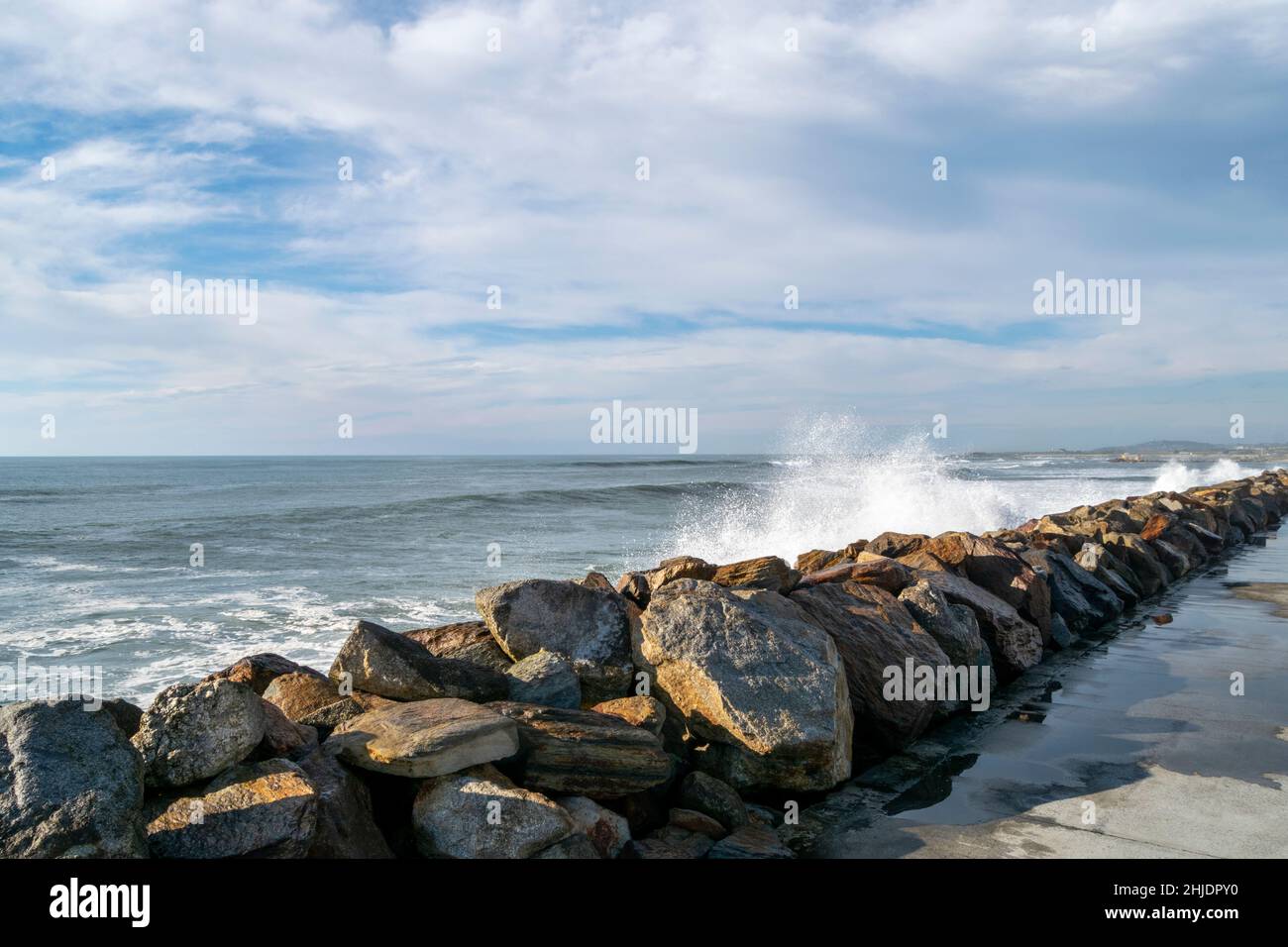Felsbarrieren Reihen sich entlang des Strandsands zu einer Meereswand mit großen Felsen, die hoch mit blauem bewölktem Himmel im Hintergrund gestapelt sind. Stockfoto