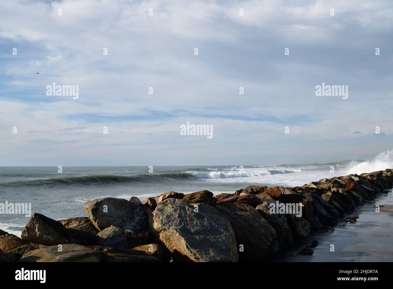 Felsbarrieren Reihen sich entlang des Strandsands zu einer Meereswand mit großen Felsen, die hoch mit blauem bewölktem Himmel im Hintergrund gestapelt sind. Stockfoto