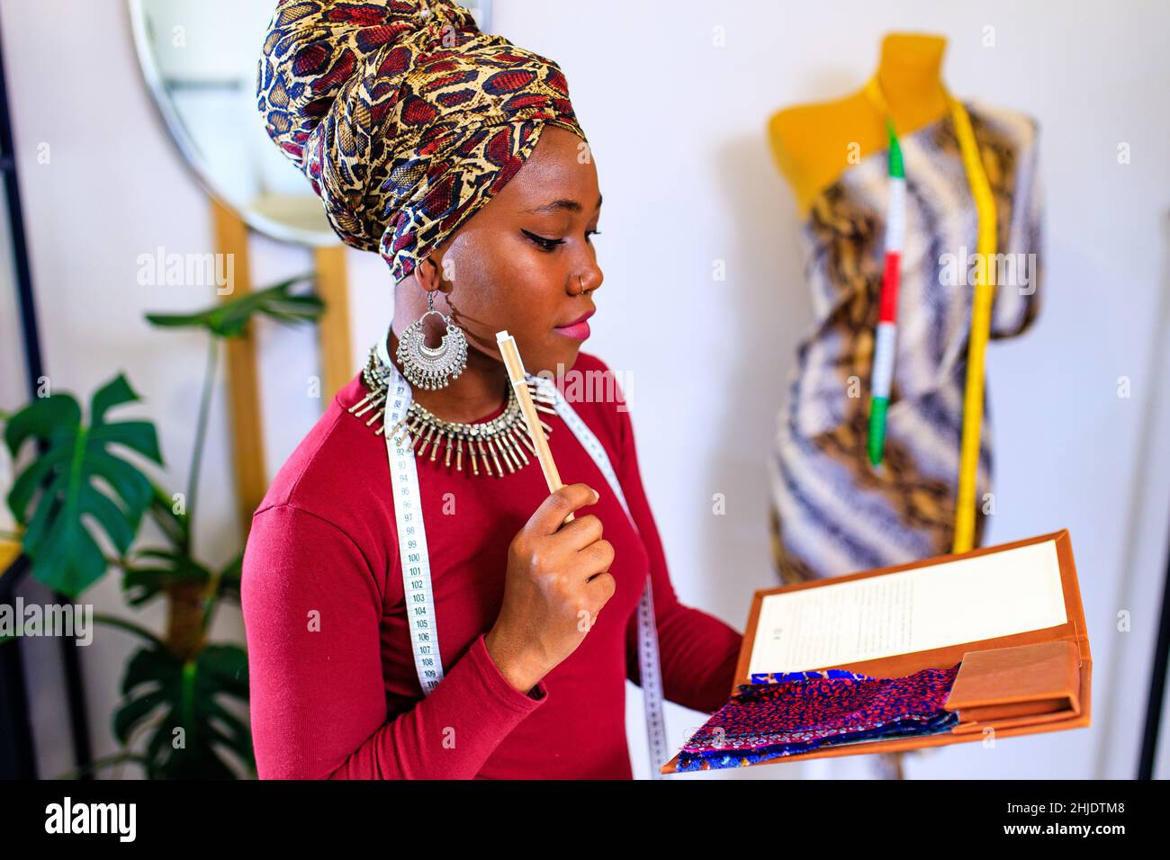 lateinamerikanische hispanische Frau mit modischem Turban über dem Kopf und rotem Kleid, die in der Kleiderherstellung im Büro arbeitet Stockfoto