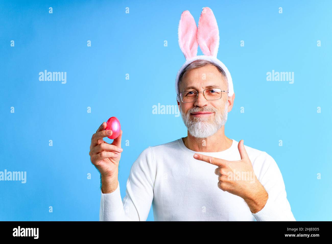 Niedlicher reifer Mann mit Hasenohren, die Osterei auf blauem Hintergrund halten Stockfoto