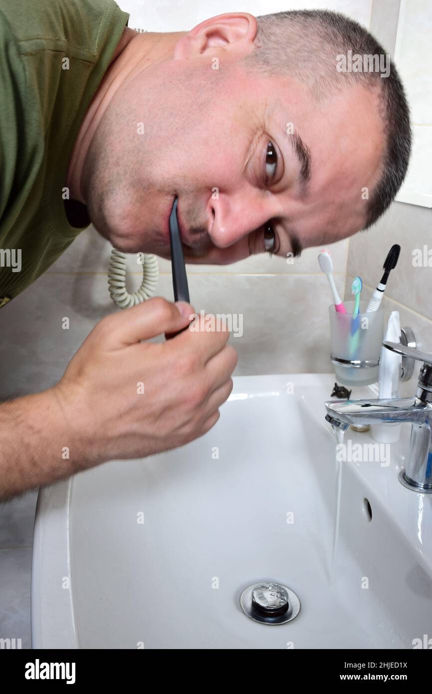 Ein weißer Mann putzt sich die Zähne in der Badewanne über dem Waschbecken. Er schaut in die Kamera. Der Wasserhahn ist offen und Wasser fließt. Stockfoto