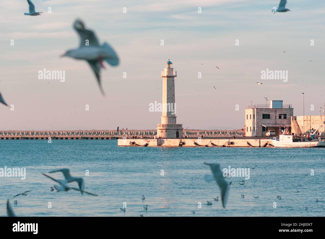 Schöner Leuchtturm mit ruhigem blauem Meer, Möwen und Fischerboot, das in der Nähe des Kais des Hafens geparkt ist Stockfoto