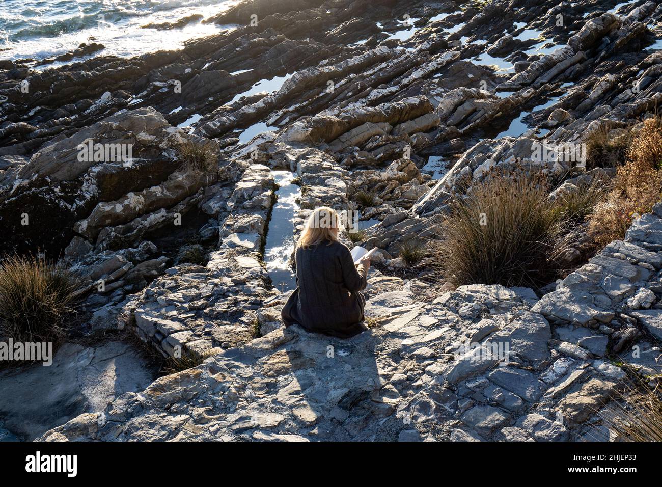 Italien, Genua - Eine junge Frau, die auf den Klippen sitzt, liest ein Buch und entspannt sich im warmen Licht des Sonnenuntergangs im alten Dorf Nervi Stockfoto