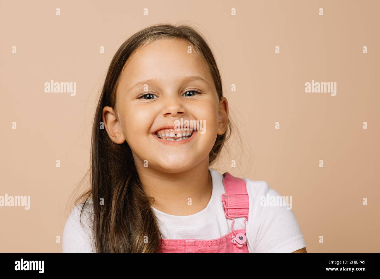 Kleines Mädchen mit leuchtend leuchtenden Augen und aufgeregt glückseligen Lächeln mit Zähnen Blick auf die Kamera in leuchtend rosa Jumpsuit und weißem T-Shirt auf beige Stockfoto