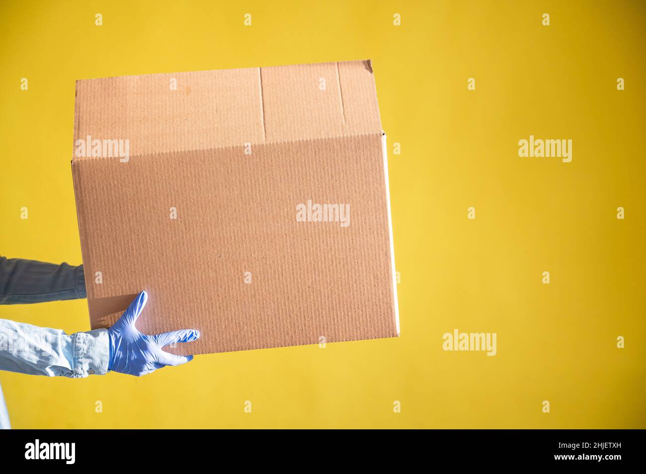 Nahaufnahme der weiblichen Hände in Handschuhen und einem Jeanshemd. Der Lieferer übergibt dem Kunden auf gelbem Hintergrund einen Karton. Antimikrobiell Stockfoto