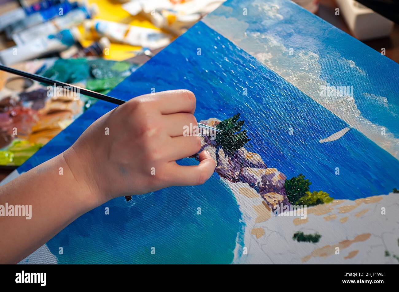 Hand des Künstlers mit einem Pinsel, der Prozess des Zeichnens von Ölgemälden mit dem Meer. Farbtuben auf unscharfem Hintergrund. Selektiver Fokus. Stockfoto