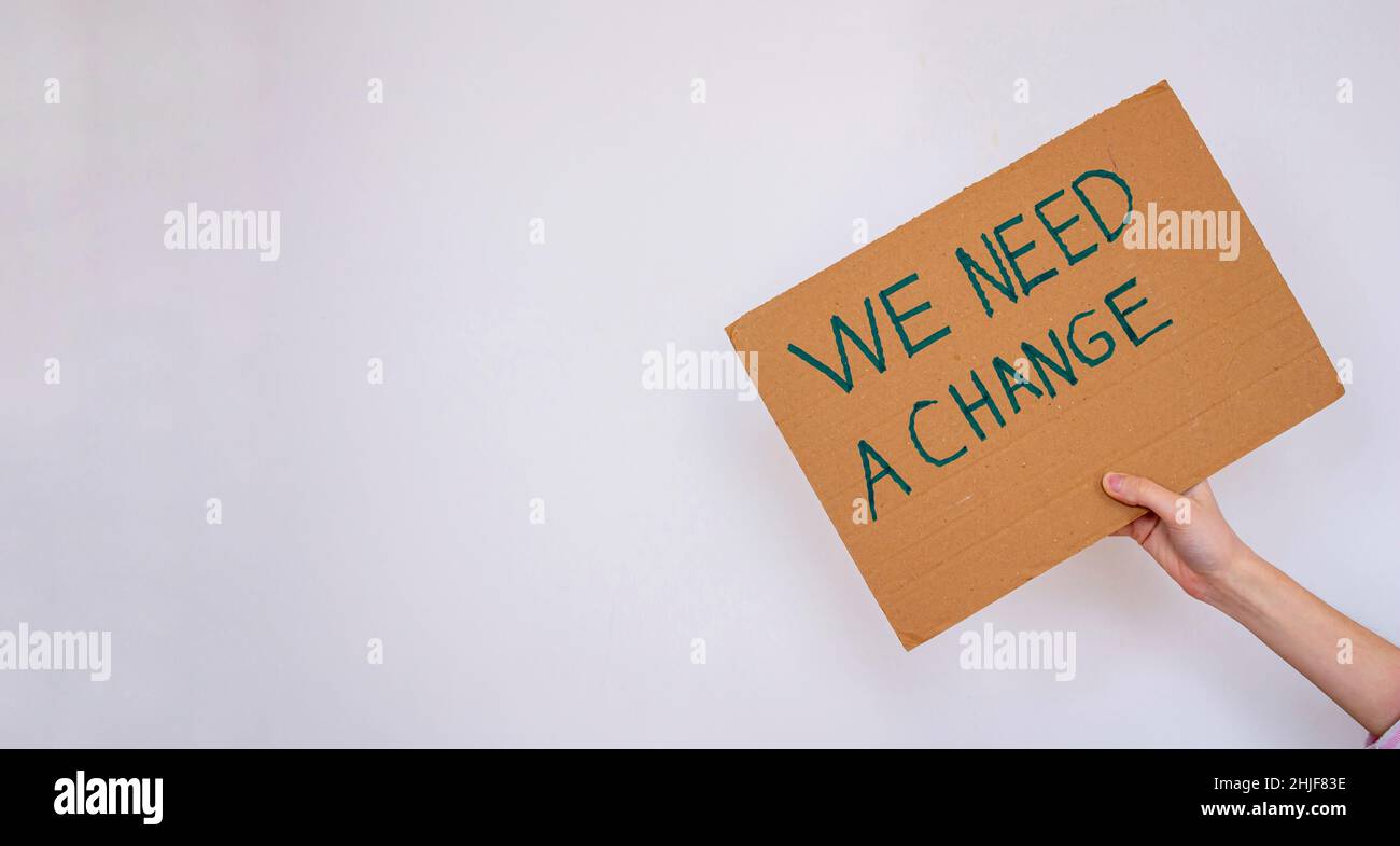 Handgehaltener Karton mit der Aufschrift „We need a change“ auf grauem Wandhintergrund. Sie kann gegen die globale Erwärmung oder Umweltverschmutzung protestieren. Stockfoto