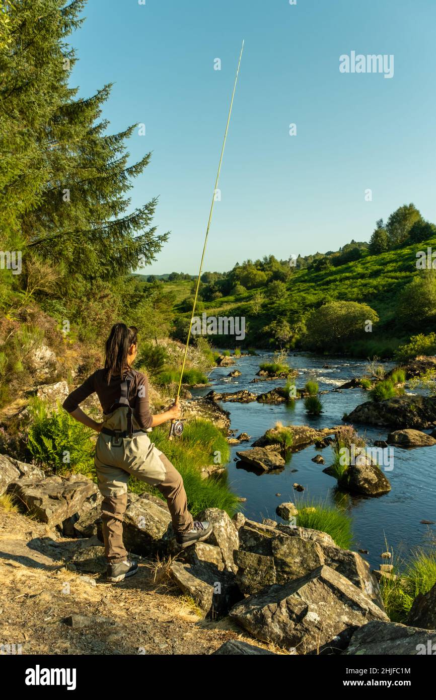 Eine asiatische weibliche Fliegenfischer Frauen, die Watvögel tragen und eine Rute halten und suchen, wo sie auf einem Fluss fischen können Stockfoto