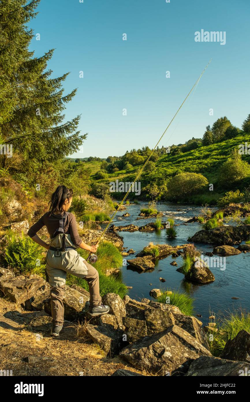 Eine asiatische weibliche Fliegenfischer Frauen, die Watvögel tragen und eine Rute halten und suchen, wo sie auf einem Fluss fischen können Stockfoto