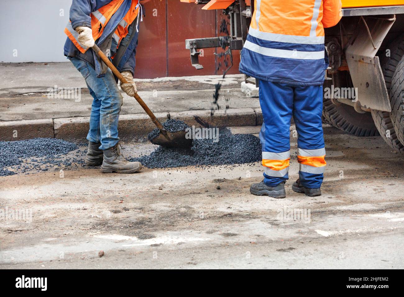 Frischer Asphalt ergießt sich auf den reparierten Straßenabschnitt in der Nähe von Arbeitern, die mit einer Schaufel in orangefarbenen Jacken in der Nähe stehen. Speicherplatz kopieren. Stockfoto