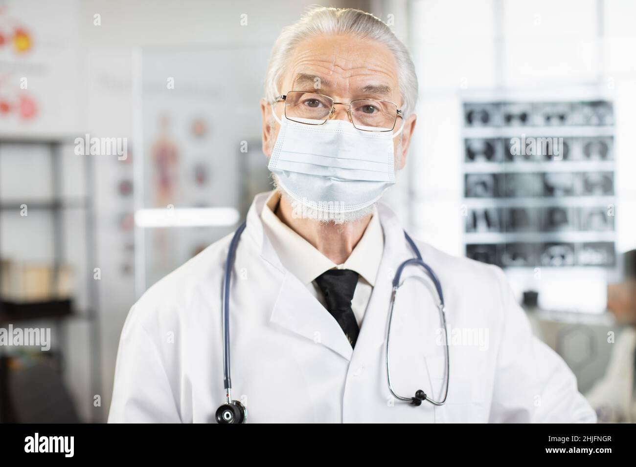 Qualifizierter Arzt im Alter mit Gesichtsmaske, Laborkittel und Brillen, die im Krankenhauszimmer stehen und die Kamera betrachten. Kaukasischer Arzt, der während des Arbeitsprozesses posiert. Stockfoto