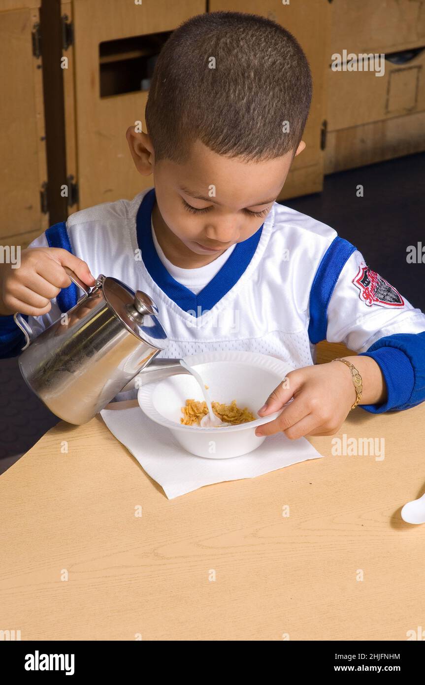 Bildung Vorschule Klassenzimmer Alter 4-5 Mahlzeit Zeit Frühstück Junge gießt Milch in seine Müslischale Stockfoto