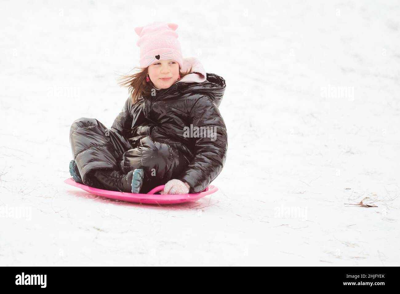 Glückliches kleines Mädchen, das auf einem Untertasse-Schlitten den Hügel hinunter rutscht. Mädchen genießen Slider Fahrt auf dem Schnee Stockfoto