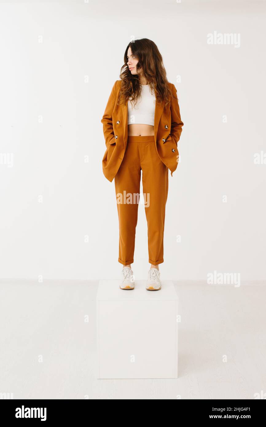 Junge Frau in modischem Pastellblazer und Hose mit stylischen Sneakers, die auf einem Würfel auf weißem Hintergrund im Studio steht. Stockfoto