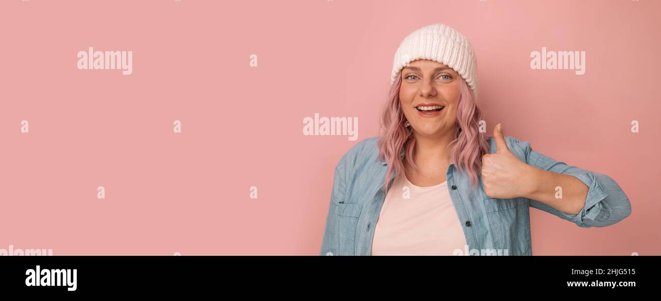 Banner zu Aufmerksamkeitsaktionen. Charmante junge kaukasische Frau in einem weißen Strickhut mit rosa Haaren zeigt Daumen nach oben wie Geste isoliert auf Pastell Stockfoto