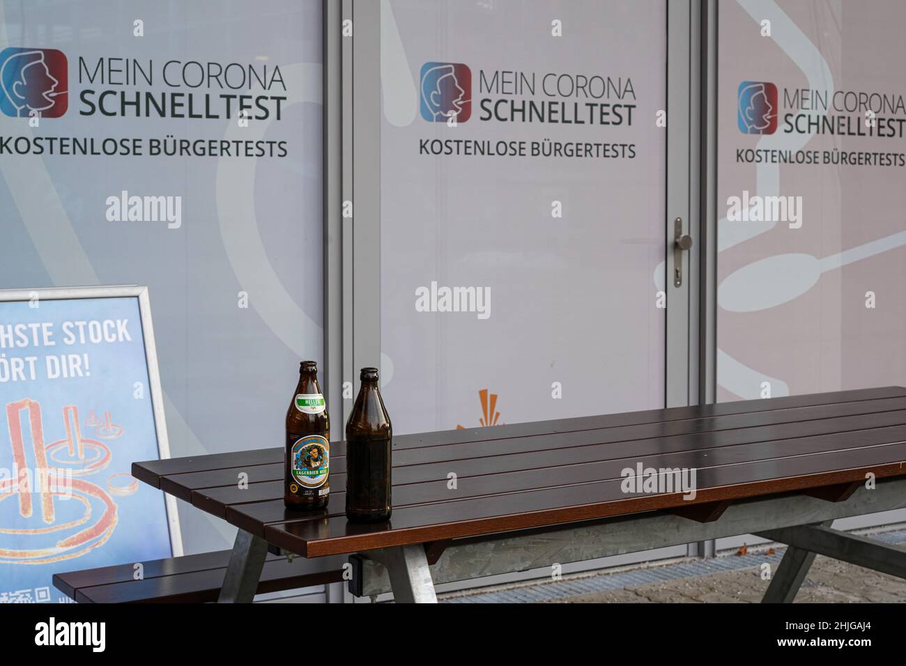 Vor der Glasscheibe eines Corona Schnelltestzentrums - Citizen Tests - befindet sich ein Holztisch und zwei Bänke. Auf dem Tisch stehen zwei leere Bierflaschen. Stockfoto