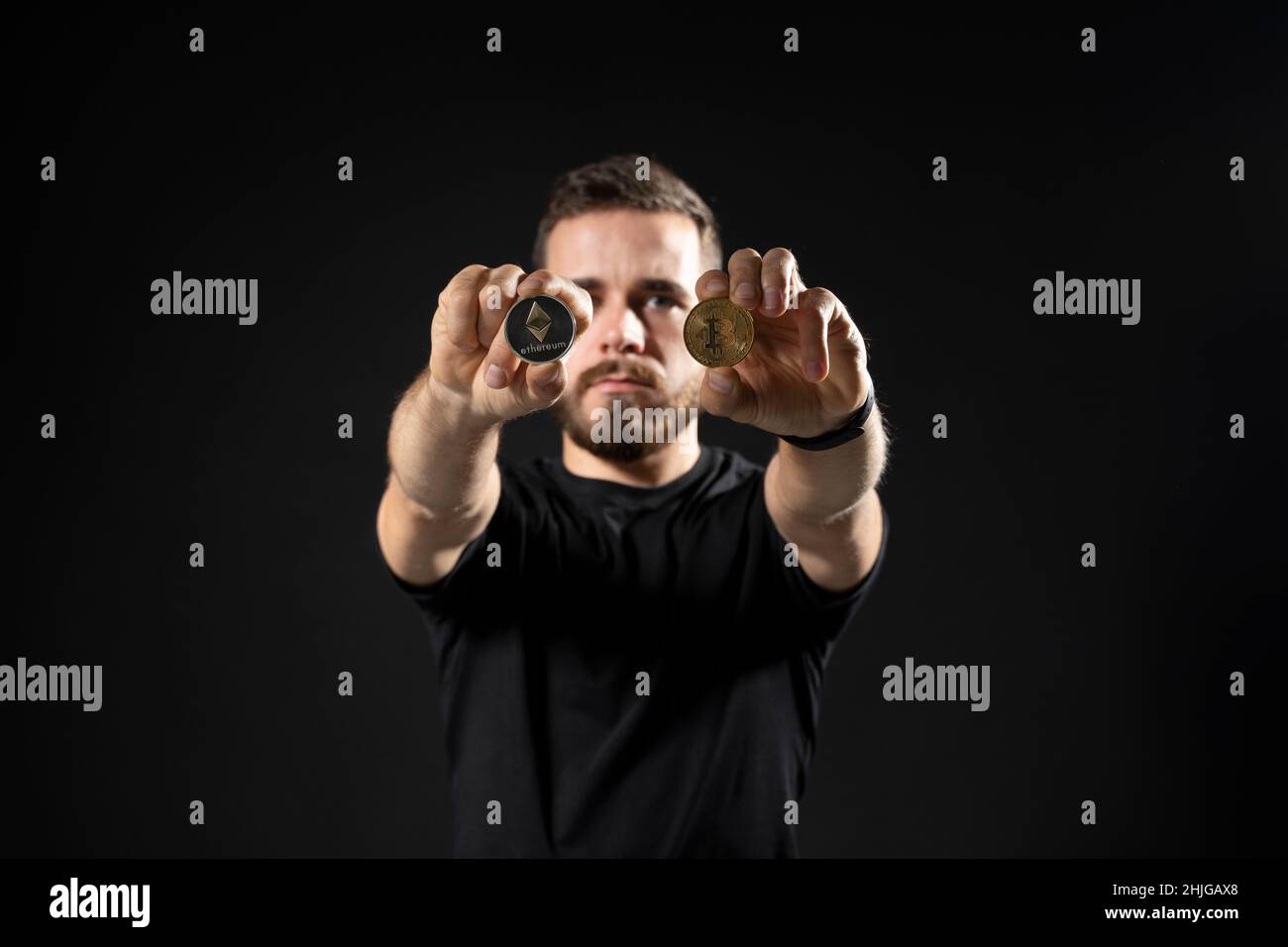 Junger bärtiger Mann mit virtueller Währung ethereum-Münze und Bitcoin auf einem schwarzen Hintergrund-Studio. Kryptowährung. Stockfoto