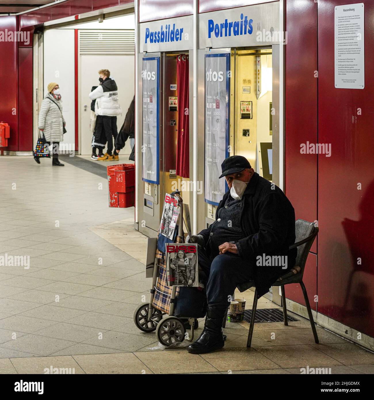 Ein Verkäufer der Straßenzeitung Biss sitzt mit Covid-Maske vor einer Fotokabine. Im Hintergrund ein junger Mann mit Maske umarmt seine Freundin Stockfoto
