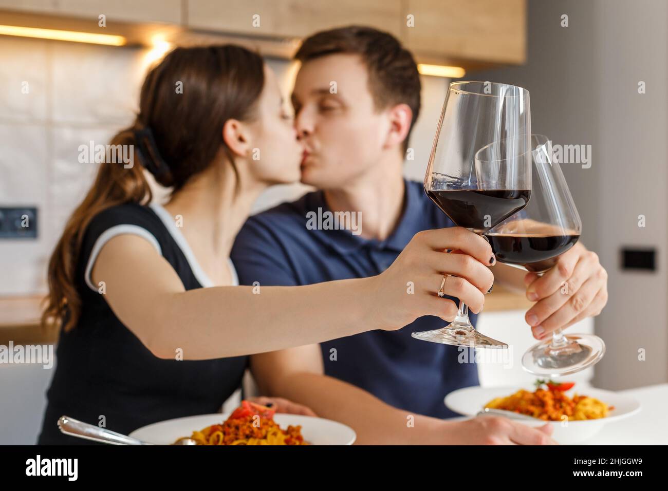 Romantisches Abendessen zu Hause. Glückliches Paar küsst, klirrende Gläser und isst Bolognese Pasta mit Wein in der Küche. Konzept von häuslicher Lebensweise, glücklicher Ehe und Zweisamkeit. Fokus auf Vordergrund. Stockfoto