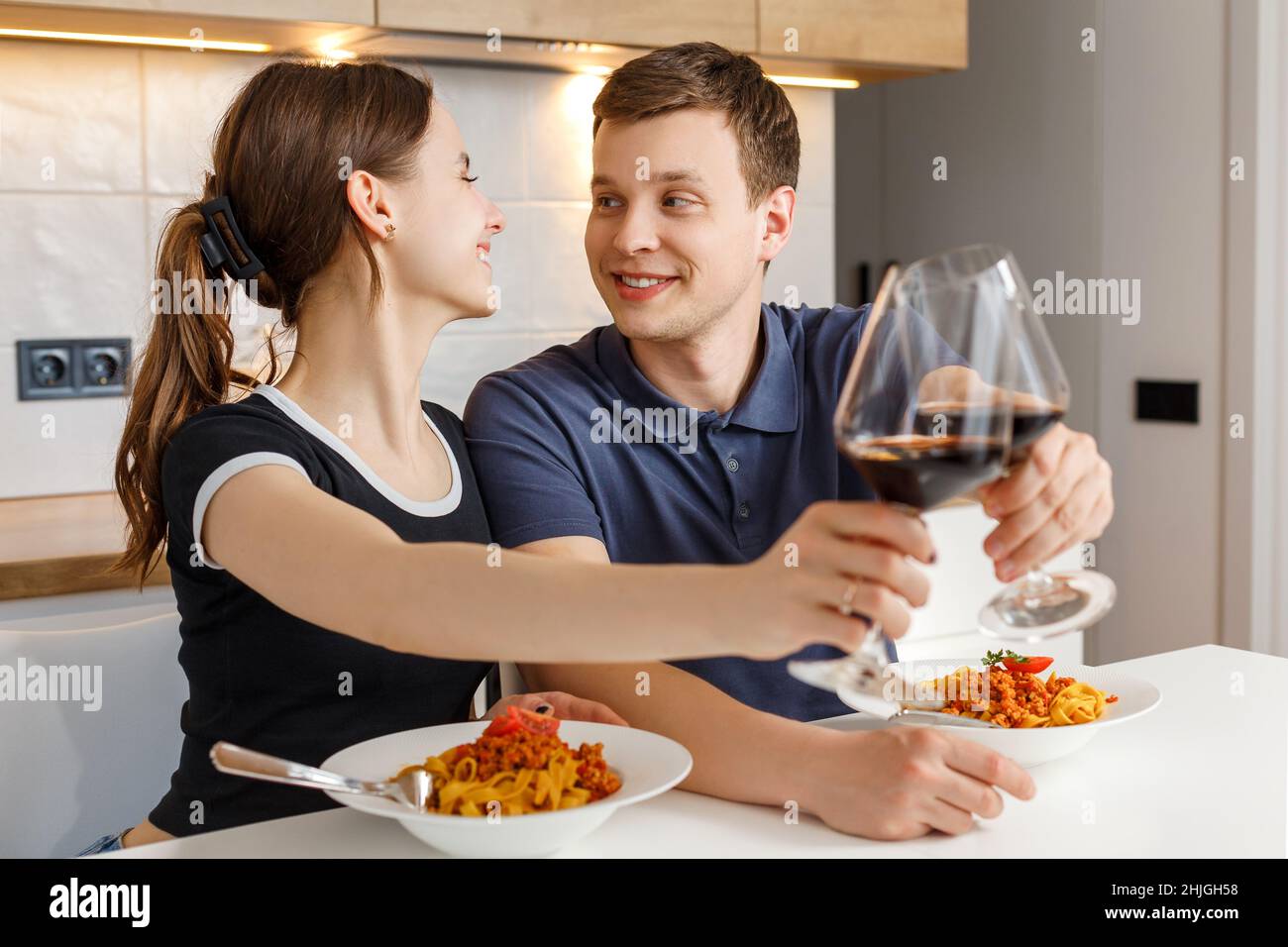 Romantisches Abendessen am Valentinstag zu Hause. Glückliches Paar lächelte, klirrende Gläser und aß Pasta mit Wein in der Küche. Konzept von häuslicher Lebensweise, Liebe, glücklicher Ehe und Zweisamkeit. Stockfoto