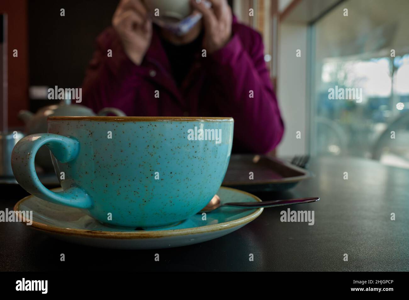 Nahaufnahme einer blauen Kaffeetasse und Untertasse mit Eierschaleneffekt auf dem Tisch im Innenbereich mit einer Person im Hintergrund, die nicht mehr scharf ist Stockfoto