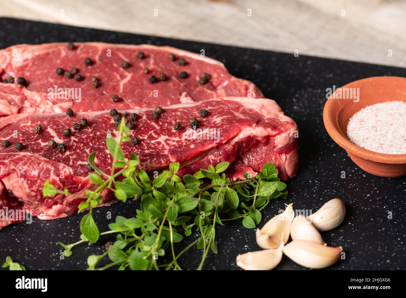 Zwei rohe, marmorierte Rindersteaks, fertig zum Kochen, mit frischem Olegano, Knoblauch und Pfefferkörnern. Stockfoto